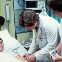 A Caucasian adult woman lying in a hospital bed, covered with a sheet. She is having blood drawn by a Caucasian male doctor with a Caucasian female nurse is in attendance. The patient is looking at the doctor and smiling.