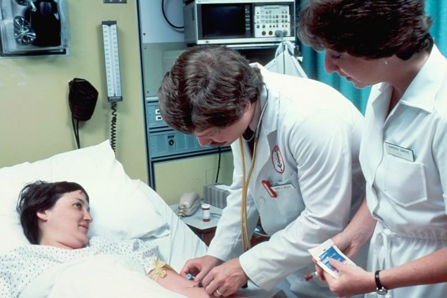 A Caucasian adult woman lying in a hospital bed, covered with a sheet. She is having blood drawn by a Caucasian male doctor with a Caucasian female nurse is in attendance. The patient is looking at the doctor and smiling.
