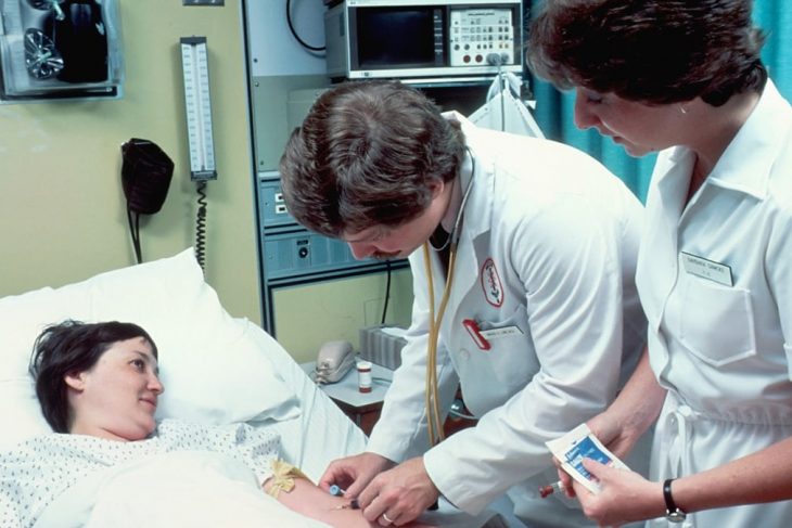 A Caucasian adult woman lying in a hospital bed, covered with a sheet. She is having blood drawn by a Caucasian male doctor with a Caucasian female nurse is in attendance. The patient is looking at the doctor and smiling.