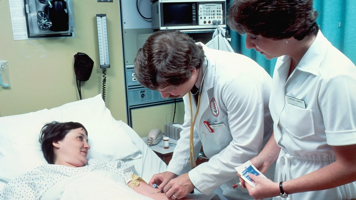A Caucasian adult woman lying in a hospital bed, covered with a sheet. She is having blood drawn by a Caucasian male doctor with a Caucasian female nurse is in attendance. The patient is looking at the doctor and smiling.